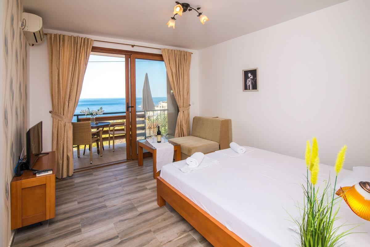 Habitació d'hotel amb llit, televisió, taula amb una ampolla i un balcó amb vista al mar.
