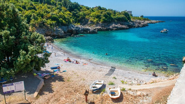 A beach with clear turquoise water, a few boats, and a few people. There are trees and buildings in the background. - Arabela 2 Apartments (Utjeha)