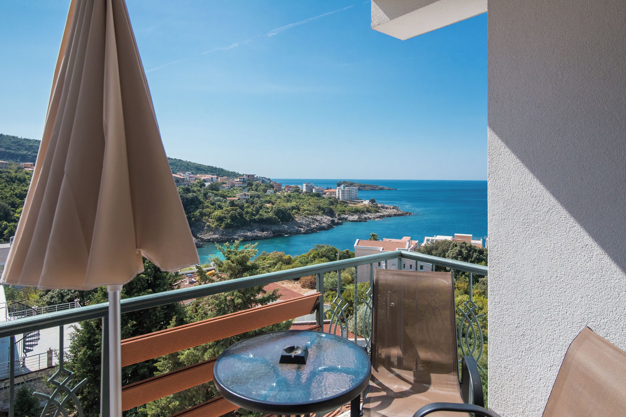 Ein Balkon mit Tisch und Stühlen, der einen Blick auf eine Küstenlandschaft mit Gebäuden und wolkenlosem Himmel bietet.
