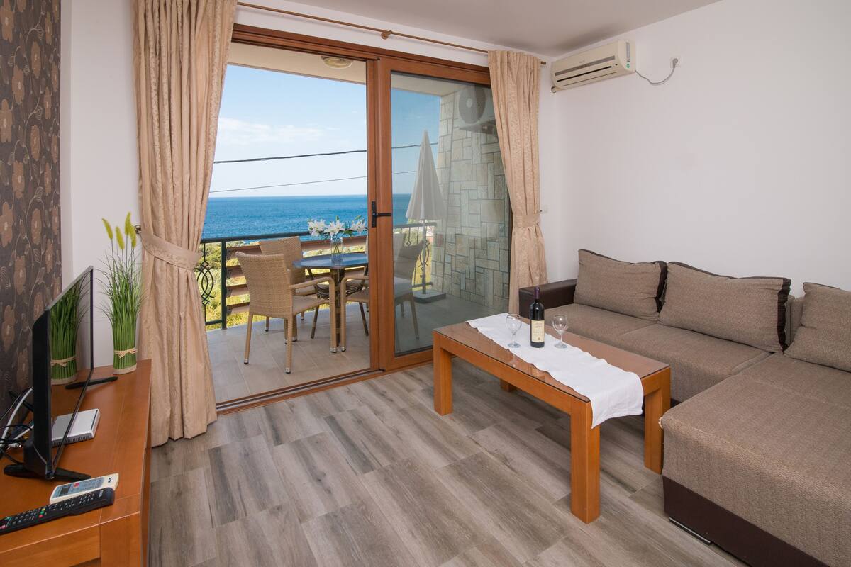 Un saló modern amb un sofà, una taula baixa i una televisió. Hi ha un balcó amb una taula i cadires, i una vista al mar.