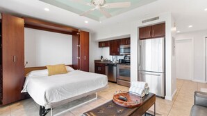 Une chambre d’hôtel moderne équipée d’un lit, d’un bureau, d’une chaise, d’une kitchenette et d’un ventilateur de plafond.