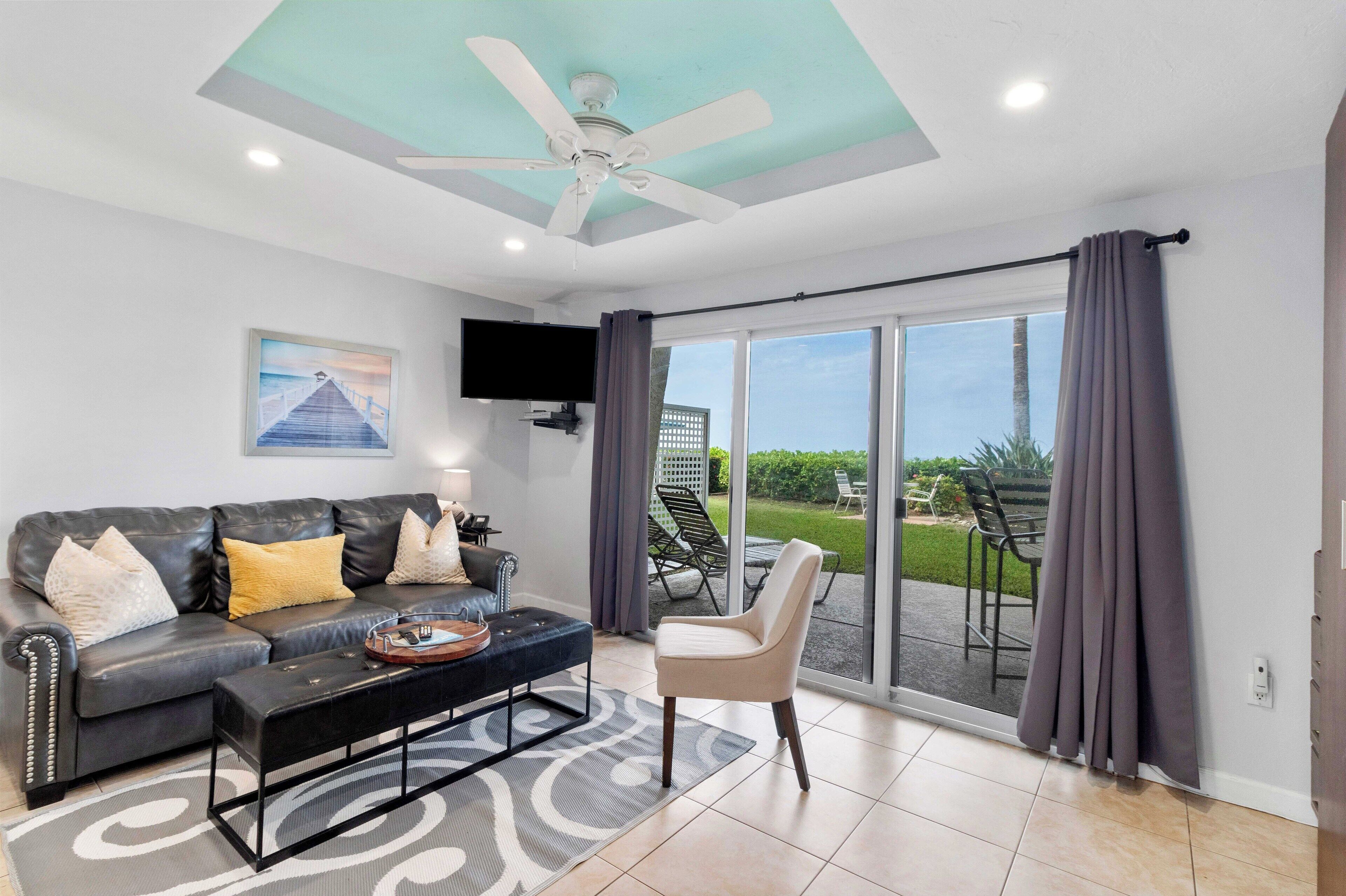 A living room with a sofa, coffee table, and a TV mounted on the wall. There is a sliding glass door leading to an outdoor area with a patio and chairs.