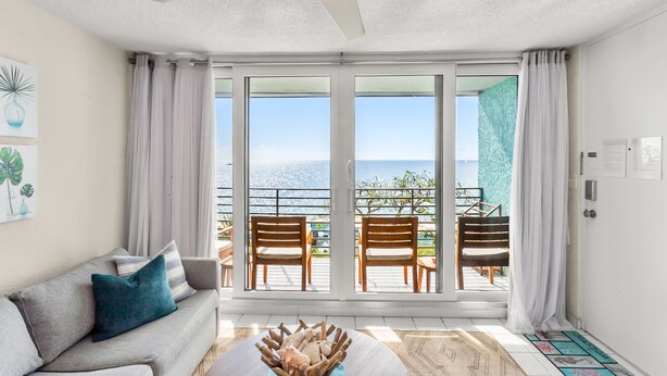 A room with a gray sofa, wooden chairs, a ceiling fan, and a view of the ocean through large sliding doors. - Ocean View, Spacious Condo next to Pool (St Thomas)