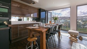 A modern kitchen with wooden cabinets, a dining area with a wooden table and chairs, and large windows offering a city view.