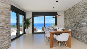 Ett modernt matområde med utsikt över havet, en stor krukväxt och ett matbord i trä med vita stolar.