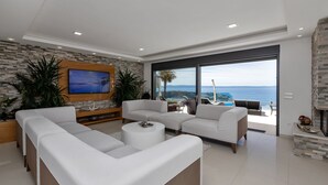 Un salon moderne doté d’un grand téléviseur, de canapés blancs, d’une table basse et offrant une vue sur la mer grâce à de grandes fenêtres.