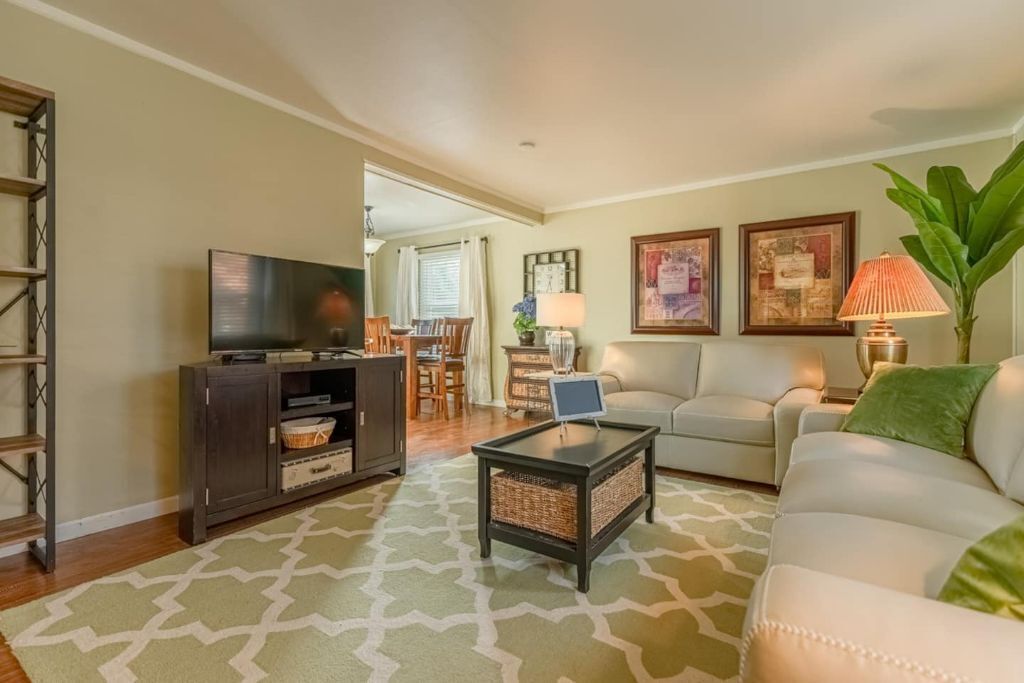 A living room with a sofa, a coffee table, a TV, and a bookshelf. There is a dining area in the background with a table and chairs.