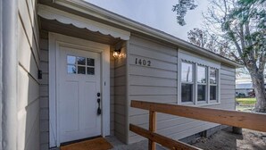A single-story house with a white exterior, numbered 1402, featuring a front door with a small window, a wooden bench outside, and a doormat. - Kimball Cottage (Richland)