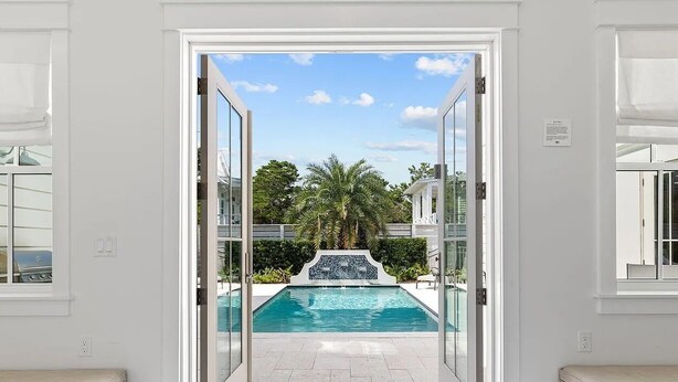 A view through a glass door leading to a swimming pool with a waterfall feature, surrounded by a white exterior and palm trees.