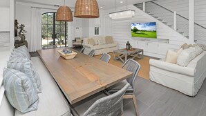 A spacious living room with a large wooden dining table, white sofas, wicker chairs, and a TV mounted on the wall.