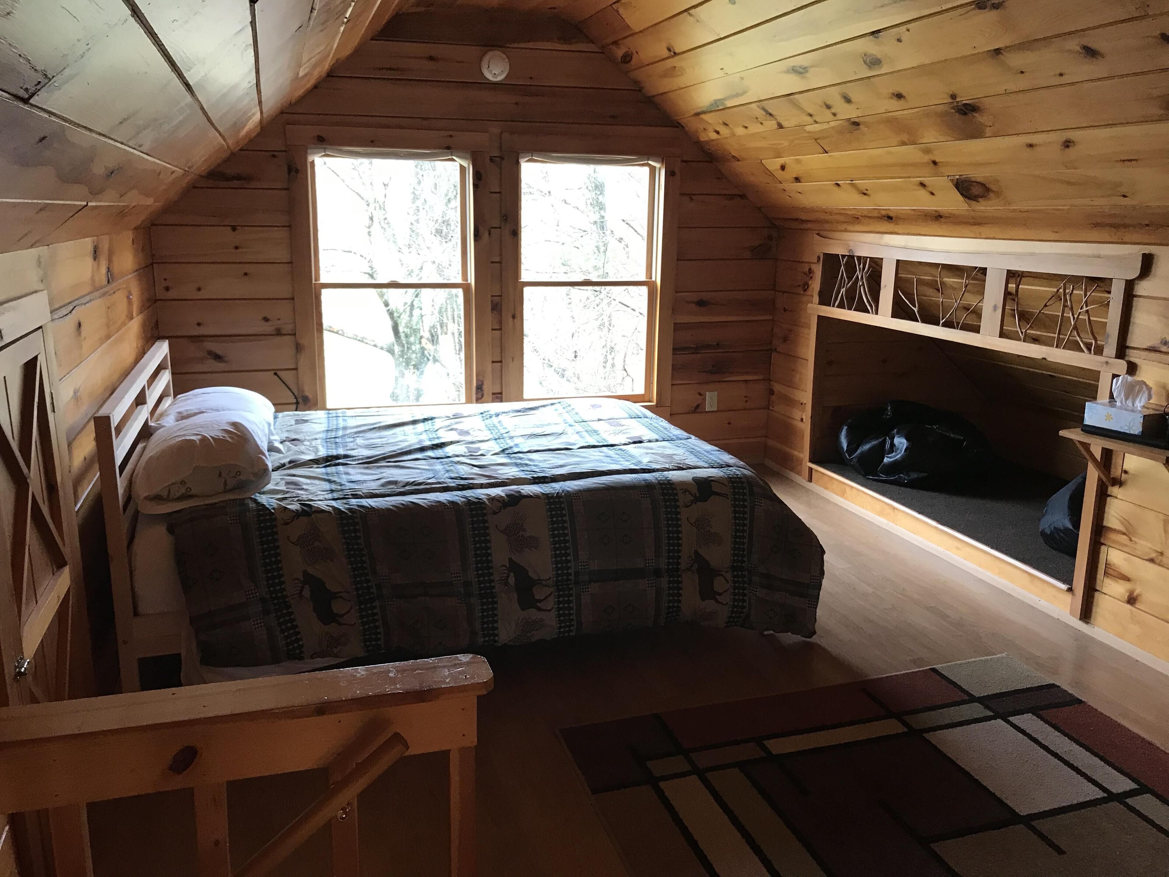 En stuga med träpanel, inredd med en säng, en bänk och ett litet bord med en näsdukslåda.
