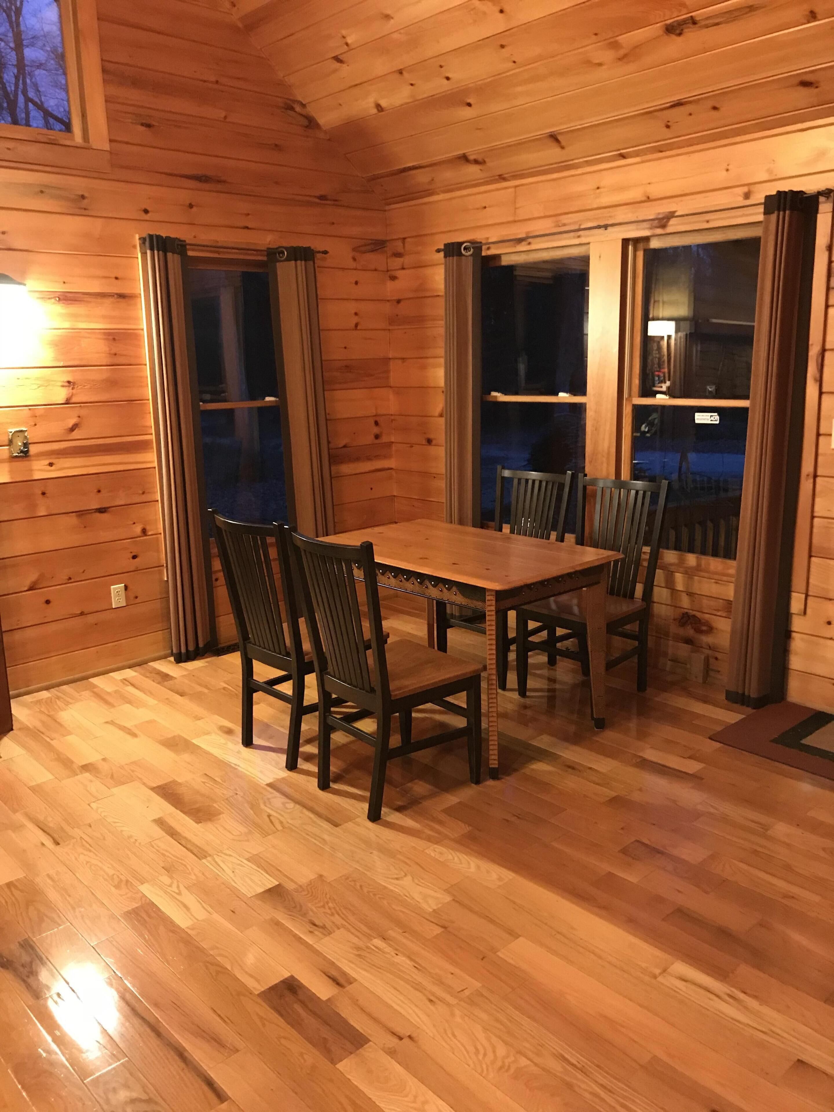 Een houten interieur met een eettafel en stoelen, grote ramen met gordijnen, en een houten vloer.