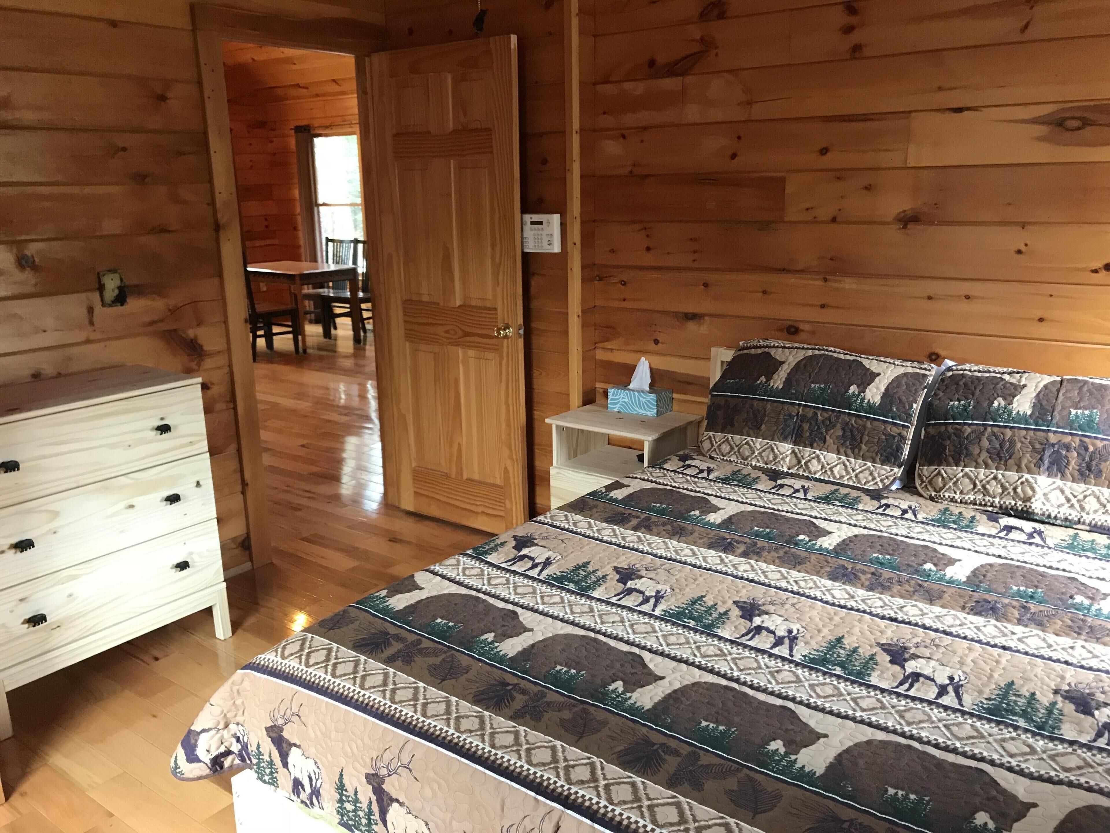Quarto de cabana de madeira com cama, cômoda, mesa de cabeceira e porta que dá para uma sala de jantar.
