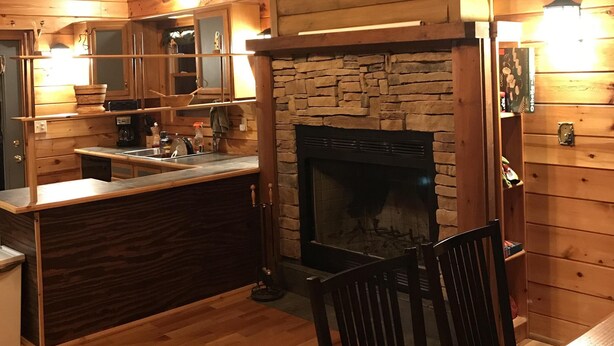 A cozy cabin interior with a stone fireplace, wooden walls, a kitchen area, and a dining table with chairs. - Cabin at Scenic Heights with a hot tub and wi-fi (Campton)