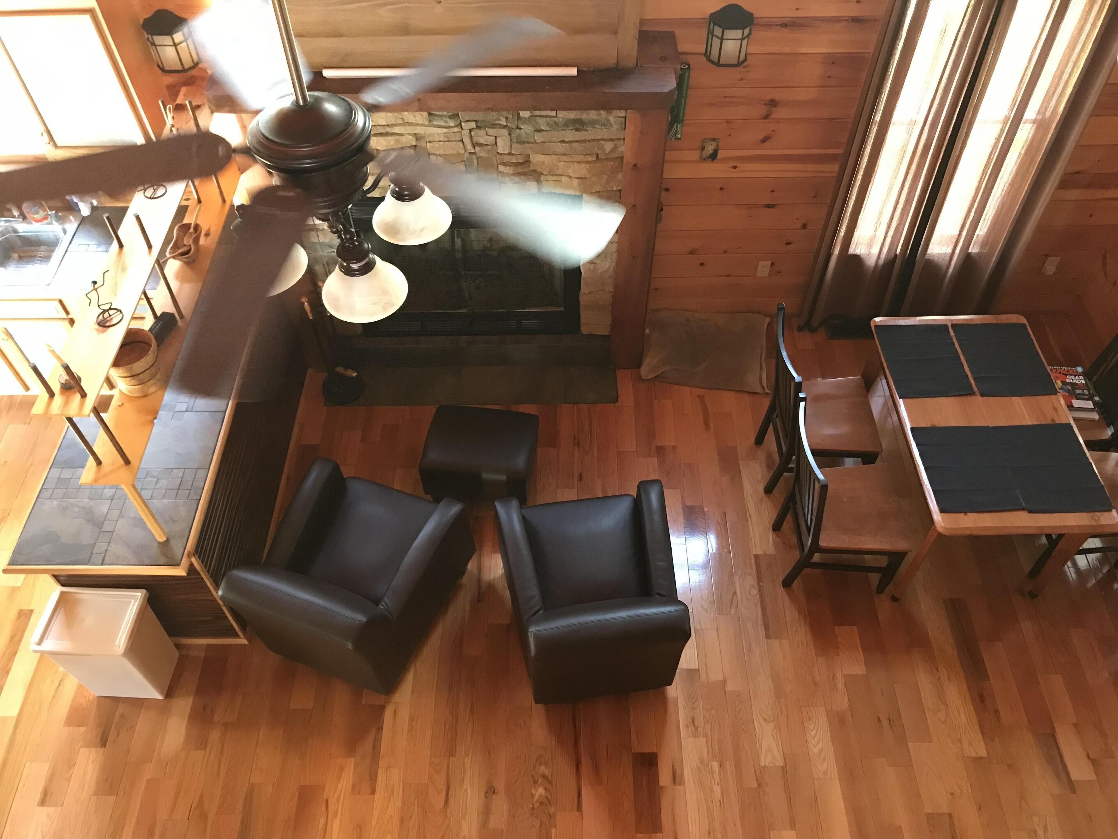 Une pièce chaleureuse avec un ventilateur de plafond, une cheminée, un plancher en bois et un coin repas comprenant des chaises et une table.
