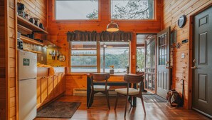 Une cuisine chaleureuse, aménagée dans le style d’un chalet, avec des murs en bois, une table à manger avec des chaises, un réfrigérateur et une grande fenêtre donnant sur une zone boisée.