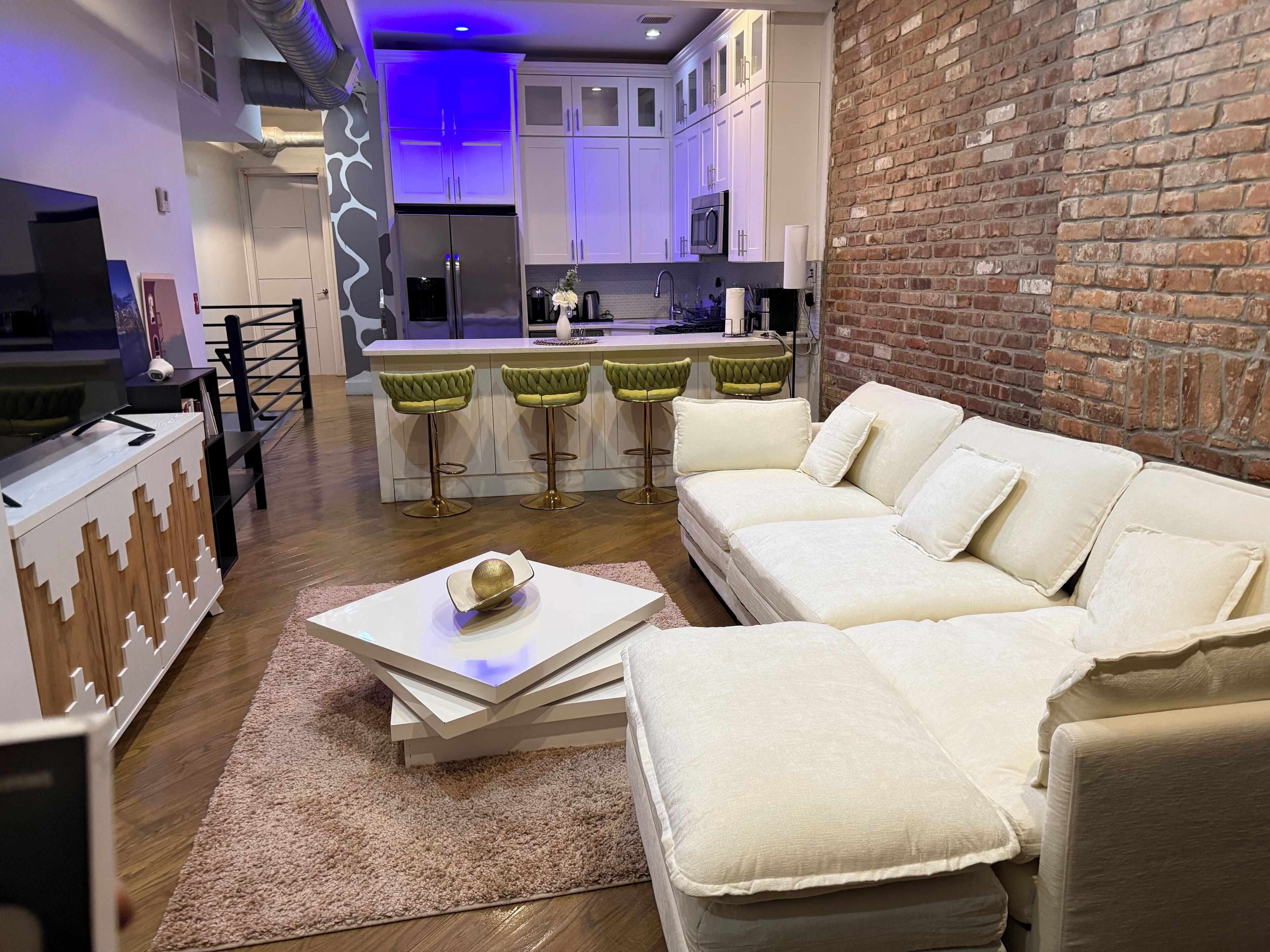 A modern living room with a white sofa, a coffee table, and a TV. There is a kitchen area with white cabinets and a brick wall.