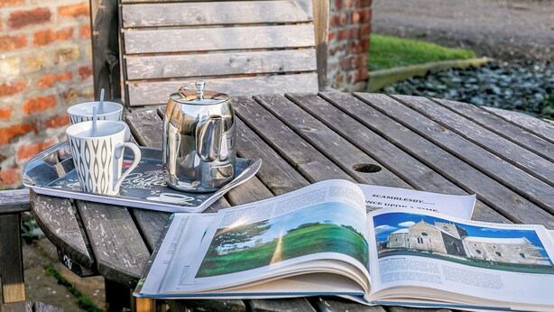 A wooden table with a metal tray holding two cups and a teapot, an open magazine, and a brick wall in the background. - Brook House Farm Cottage (Scamblesby, near Louth)