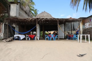 Una cabaña en la playa con techo de paja, mobiliario exterior con sillas de plástico y una hamaca.