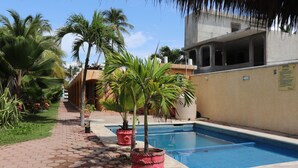 Una piscina con borde de azulejos, rodeada por un camino de ladrillo, palmeras y un edificio con un letrero que indica un hotel.