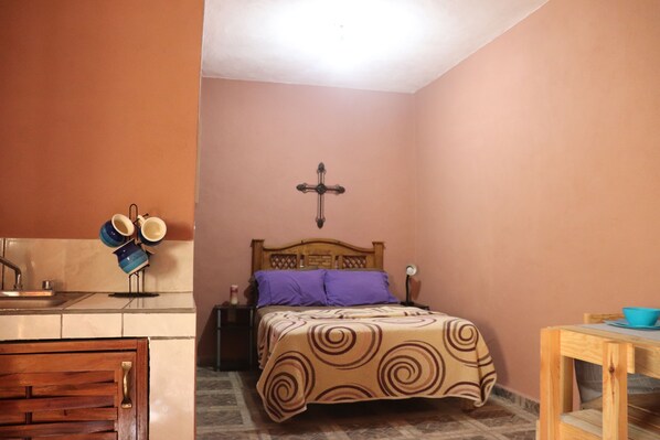 Un dormitorio con cama, mesita de noche, cabecera de madera, una cruz en la pared, una pequeña mesa con un cuenco y una zona de cocina con fregadero y utensilios.