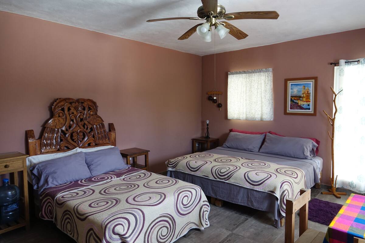 A bedroom with two beds, a wooden headboard, a ceiling fan, a water dispenser, and a window with curtains.