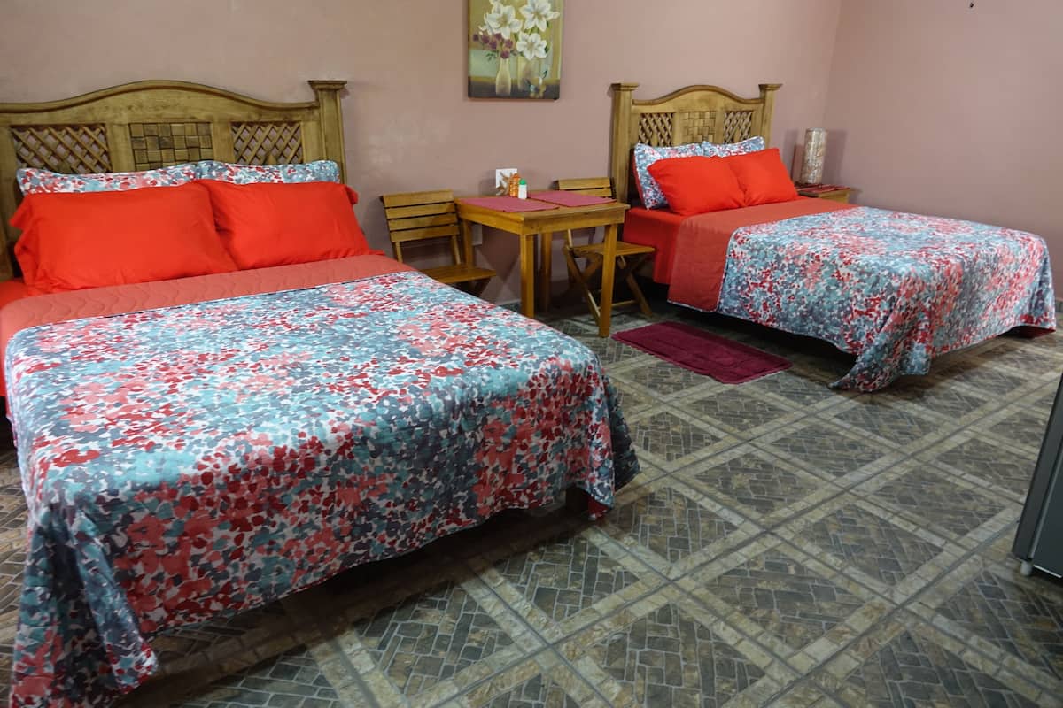 A room with two beds, each with a floral bedspread, a wooden headboard, and red pillows. There is a small table with a chair, a red rug, and a painting on the wall.