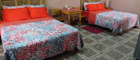 Habitación con dos camas, cada una con cubrecama floral, cabecero de madera y almohadas rojas. Hay una mesita con una silla, un tapete rojo y un cuadro en la pared.