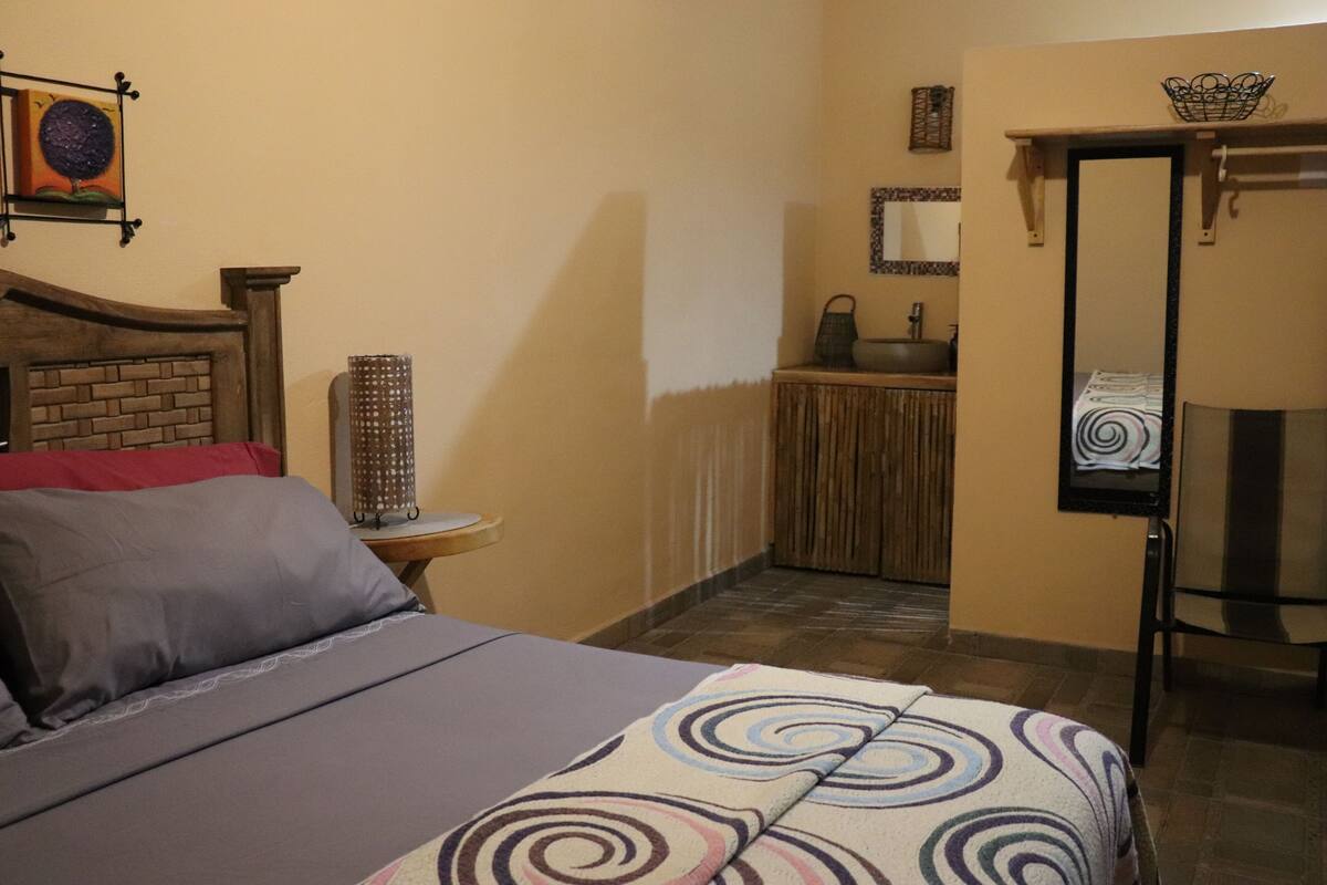 A bedroom with a bed, a nightstand, a mirror, a door, and a small table with a lamp.