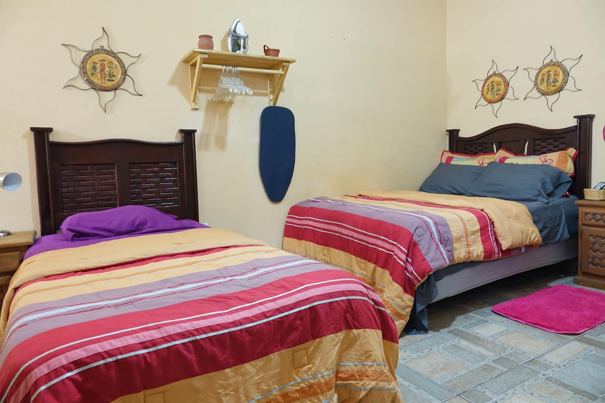 A room with two beds, each with striped bedding, a wooden headboard, and a nightstand with a lamp. There are decorative wall hangings and a shelf with items on the wall.