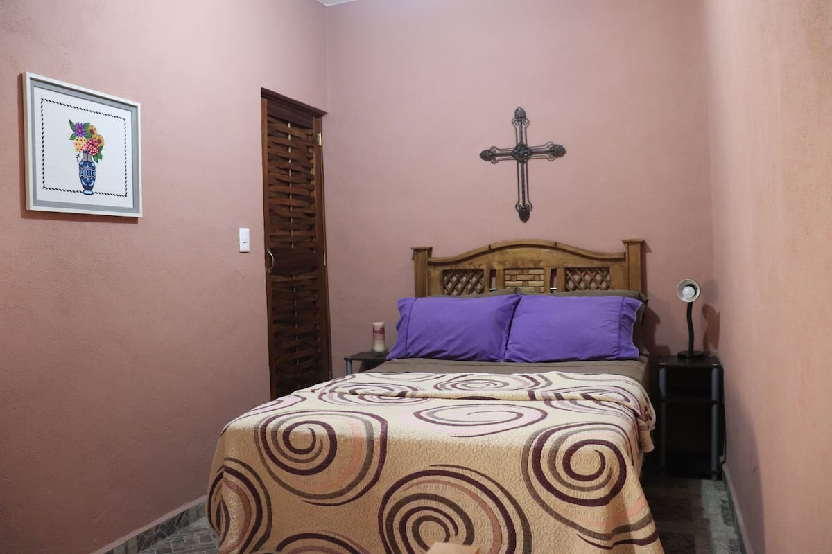 A bedroom with a bed, a wooden headboard, a purple pillow, a bedside lamp, and a framed picture on the wall.