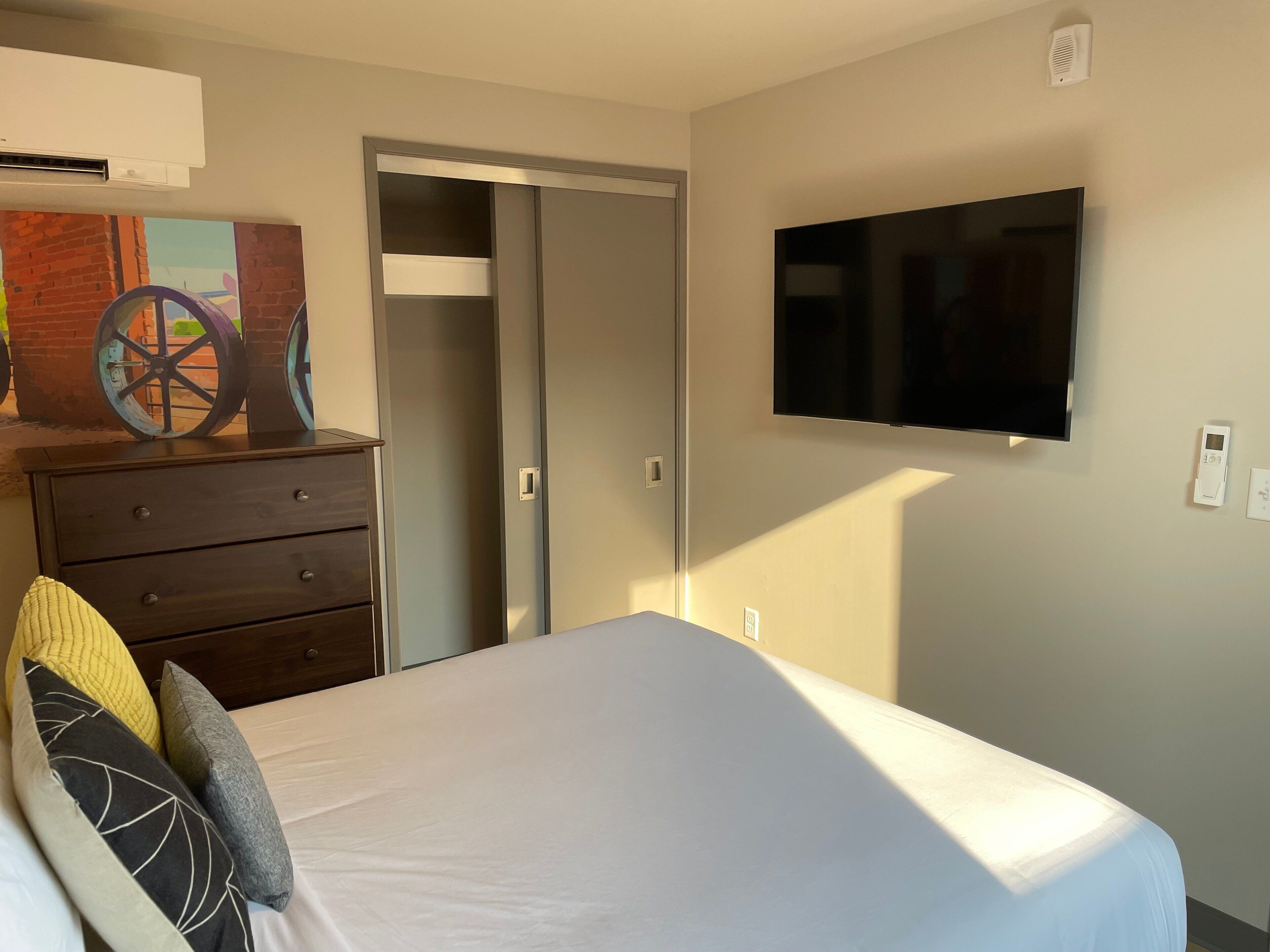 A bedroom with a bed, a dresser, a TV mounted on the wall, and a door leading to another room.