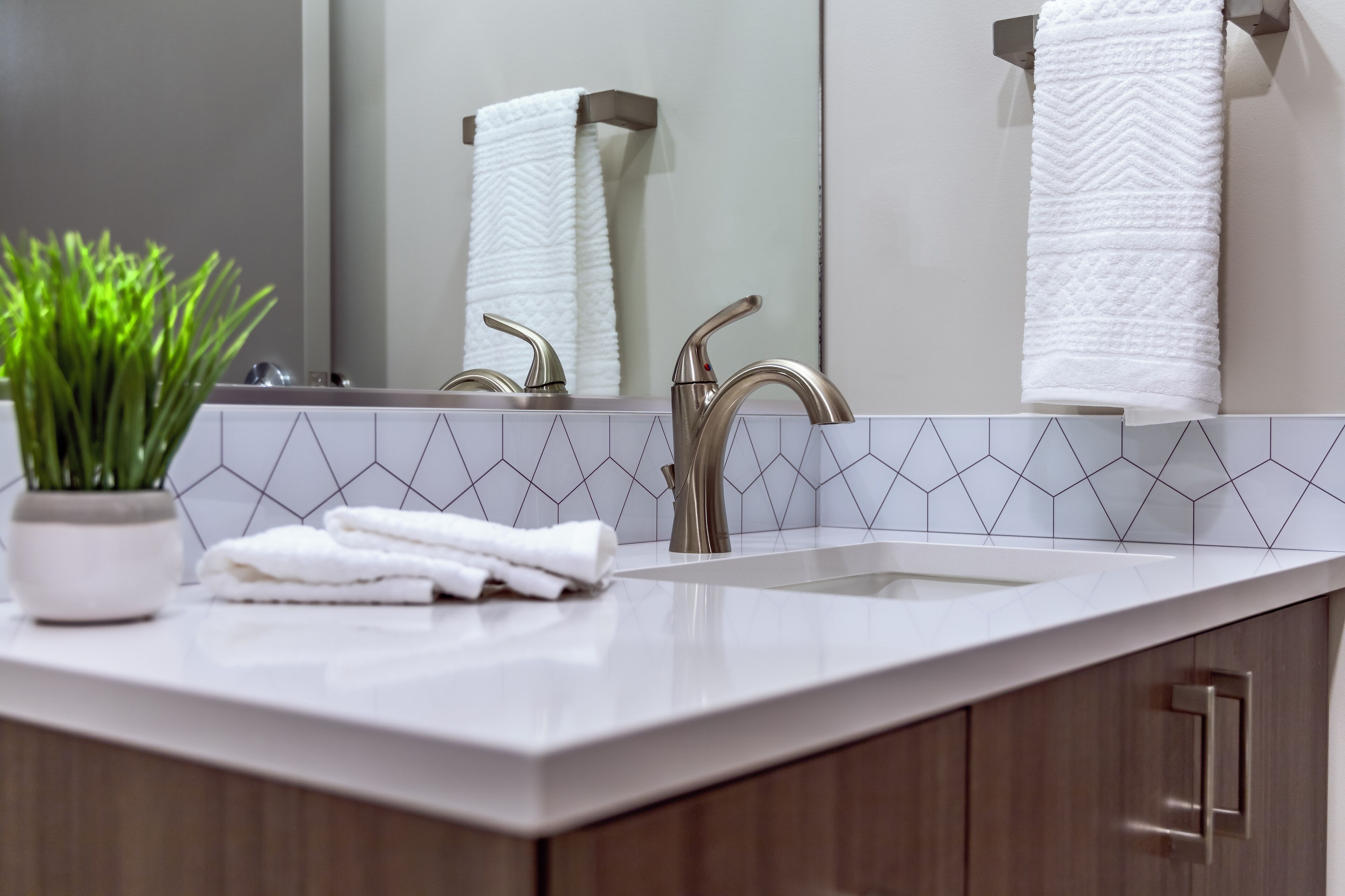 A modern bathroom with a white countertop, two chrome faucets, a white towel, and a potted plant.