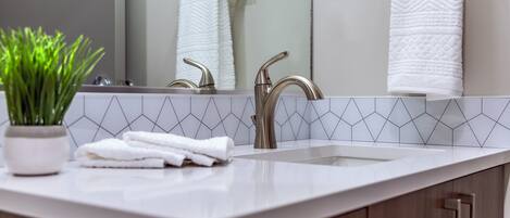 A modern bathroom with a white countertop, two chrome faucets, a white towel, and a potted plant.
