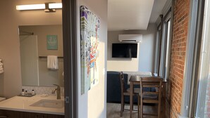 Ein modernes Badezimmer mit Waschbecken, Spiegel und Handtuchhalter. Ein farbenfrohes Kunstwerk hängt an der Wand, und ein Fernseher ist über einer Couch angebracht.