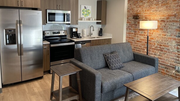 A modern kitchen with stainless steel appliances, a grey sofa, and a brick wall.