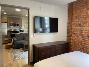 A modern hotel room with a flat-screen TV mounted on the wall, a wooden dresser, a bed with a white comforter, and a kitchenette area visible through an open door. - Attractive 1 Bed Travel Apartment Steps Away From The Spokane Opera House (Spokane)