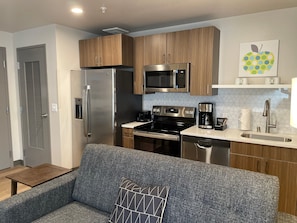 A modern kitchen with stainless steel appliances, wooden cabinets, and a gray sofa with a geometric pillow. - Attractive 1 Bed Travel Apartment Steps Away From The Spokane Opera House (Spokane)