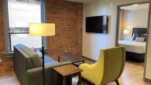 Une chambre d’hôtel moderne avec un mur en briques, un téléviseur à écran plat, un canapé, un fauteuil jaune et une petite table en bois.