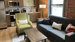 A modern kitchen with a microwave, stove, and coffee maker. There is a green armchair and a gray sofa with pillows. A lamp is on a side table.