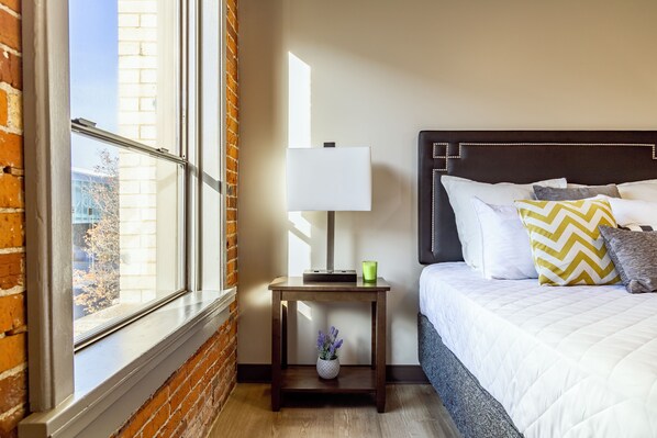 Habitación de hotel con cama, mesita de noche con lámpara, un jarrón con flores y ventana con vista a un edificio.