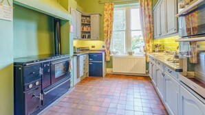 Une cuisine aux murs verts, avec un poêle d’époque et une fenêtre agrémentée de rideaux.