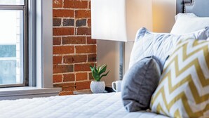 Een netjes opgemaakt bed met een witte quilt, een grijs kussen met een ruitpatroon, een witte mok en een kleine potplant op een nachtkastje.