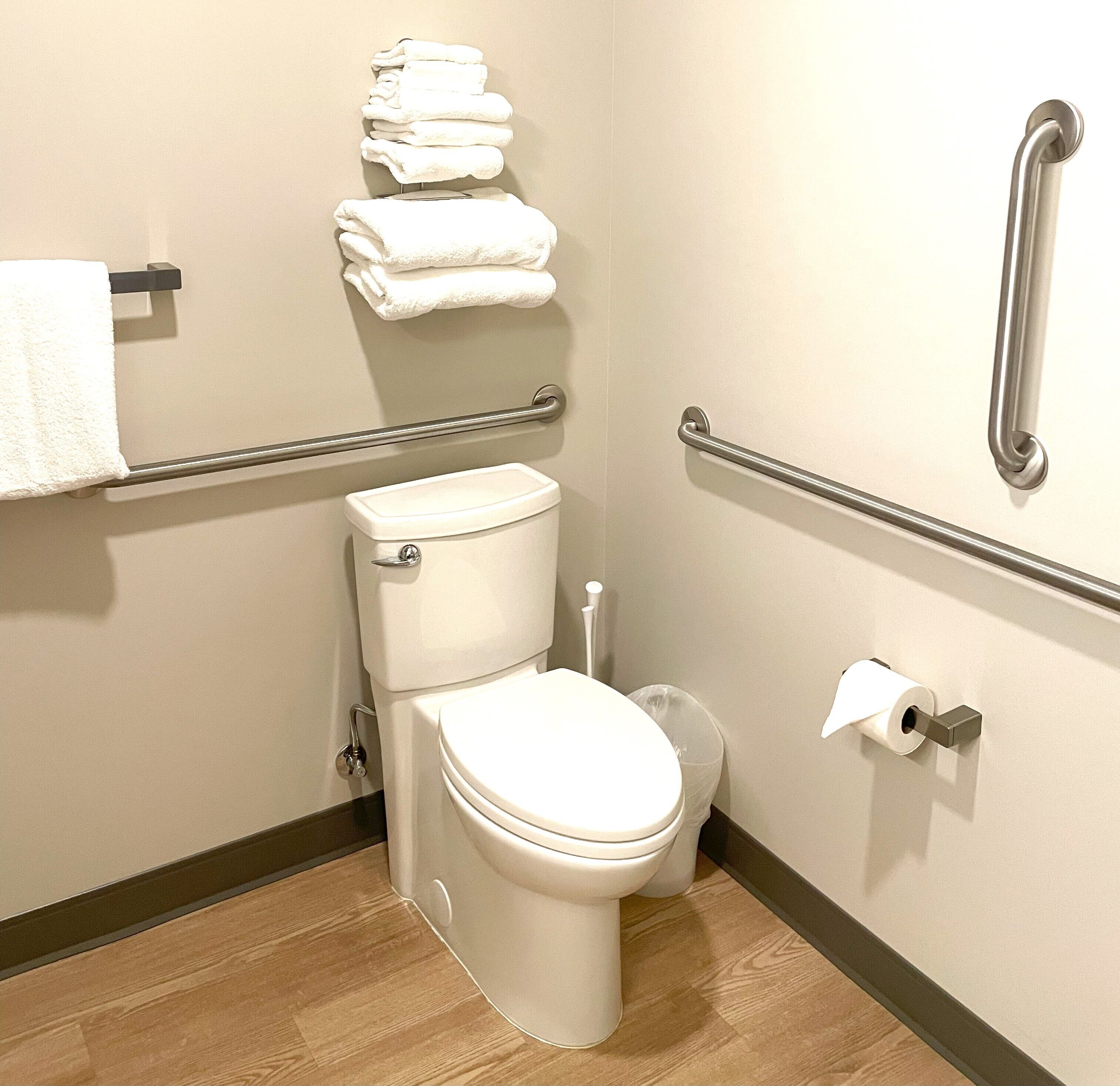 A small, modern bathroom with a toilet, a handrail, a stack of white towels, and a toilet paper holder.