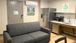 A compact living space with a grey sofa, a kitchenette with a refrigerator, microwave, and coffee maker, and a door with a security lock.