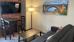 Un salon avec un téléviseur fixé à un mur en briques, une table à manger avec des chaises, un lampadaire, un canapé et un tableau représentant un pont.