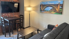Un salon avec un téléviseur fixé à un mur en briques, une table à manger avec des chaises, un lampadaire, un canapé et un tableau représentant un pont.