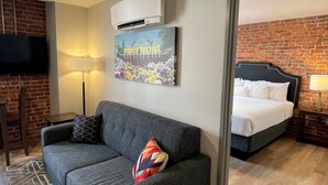 A hotel room with a grey sofa, a colorful pillow, a flat-screen TV, a painting, a bed with white bedding, and a brick wall.