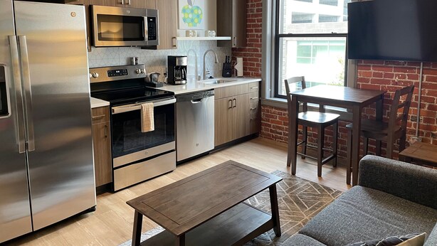 A modern kitchen with stainless steel appliances, a dining area with a table and chairs, and a large window with a brick wall.