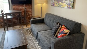 A living room with a gray sofa, a TV mounted on a brick wall, a table with chairs, and a lamp.